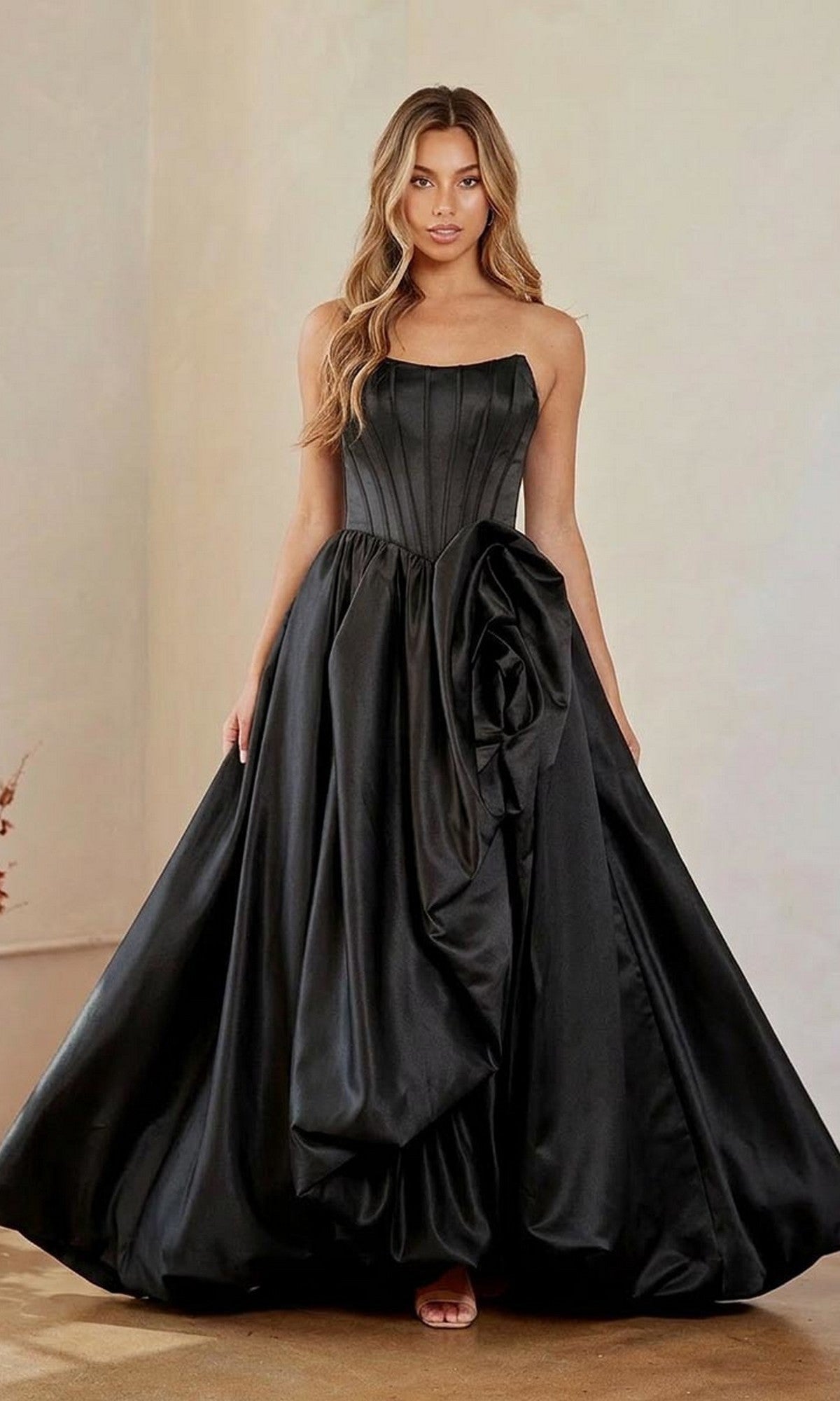 A woman stands indoors against a plain background, wearing the Long Prom Dress: Amelia Couture 2085 in black—a strapless, full-length ball gown featuring a fitted corset bodice, bubble hem, and large floral detail on the skirt.